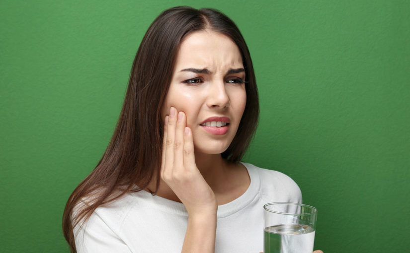 Woman with a glass of water holding her mouth and grimacing from sensitive teeth pain.